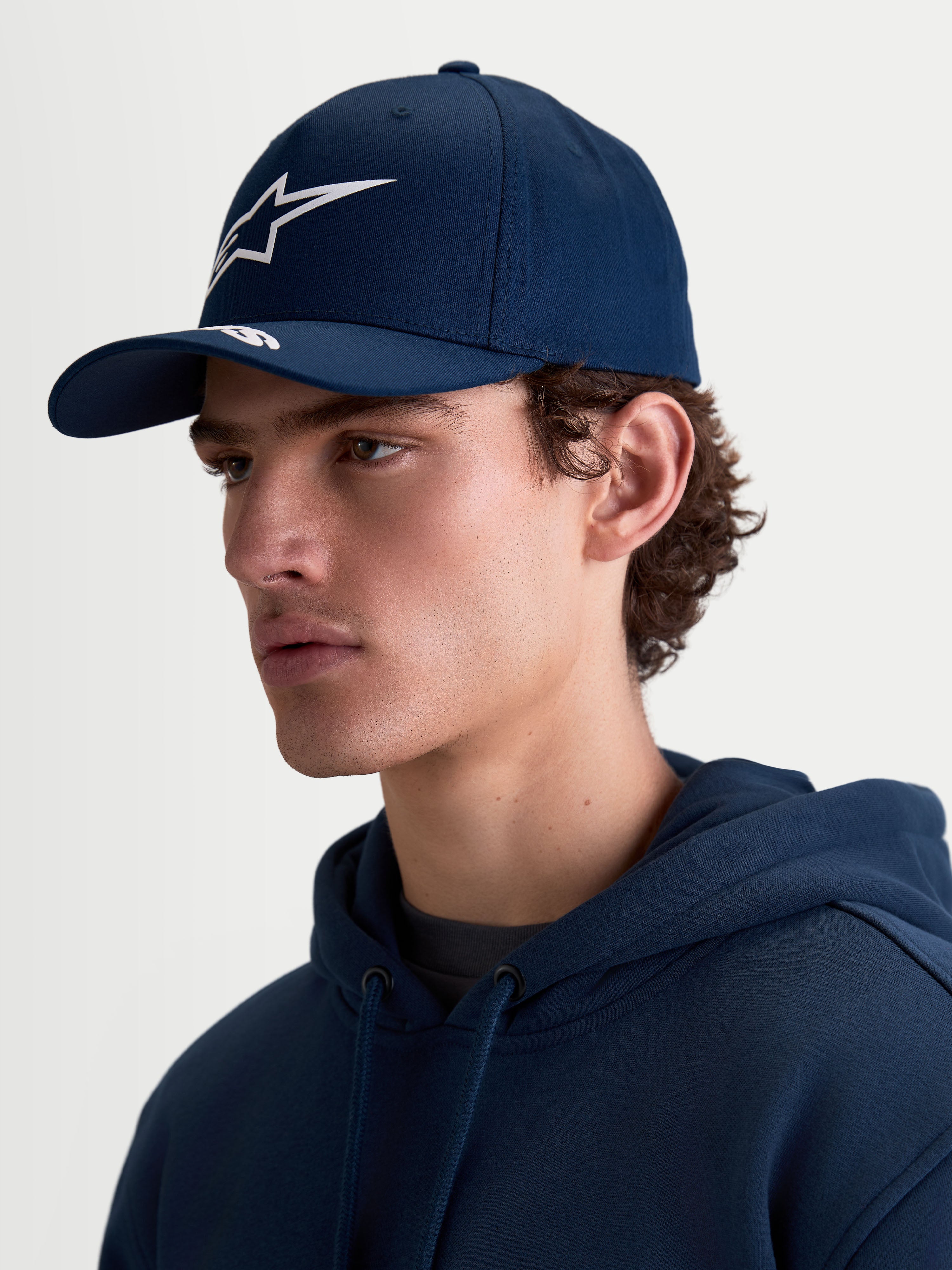 Alpinestars Arclight Hat, granatowa czapka z daszkiem typu sportswear dla kobiet i mężczyzn, z zakrzywionym daszkiem, białym logotypem Astars w technice puff print i usztywnioną koroną. Widok z profilu na modelu prezentuje klasyczny krój inspirowany motorsportem oraz trwałą tkaninę keprową.