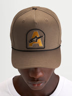 Alpinestars Czapka Acme Snapback, brązowa czapka z daszkiem typu sportswear z naszywką z haftowanym logo Astars i czarnym sznurkiem, prezentowana na modelu