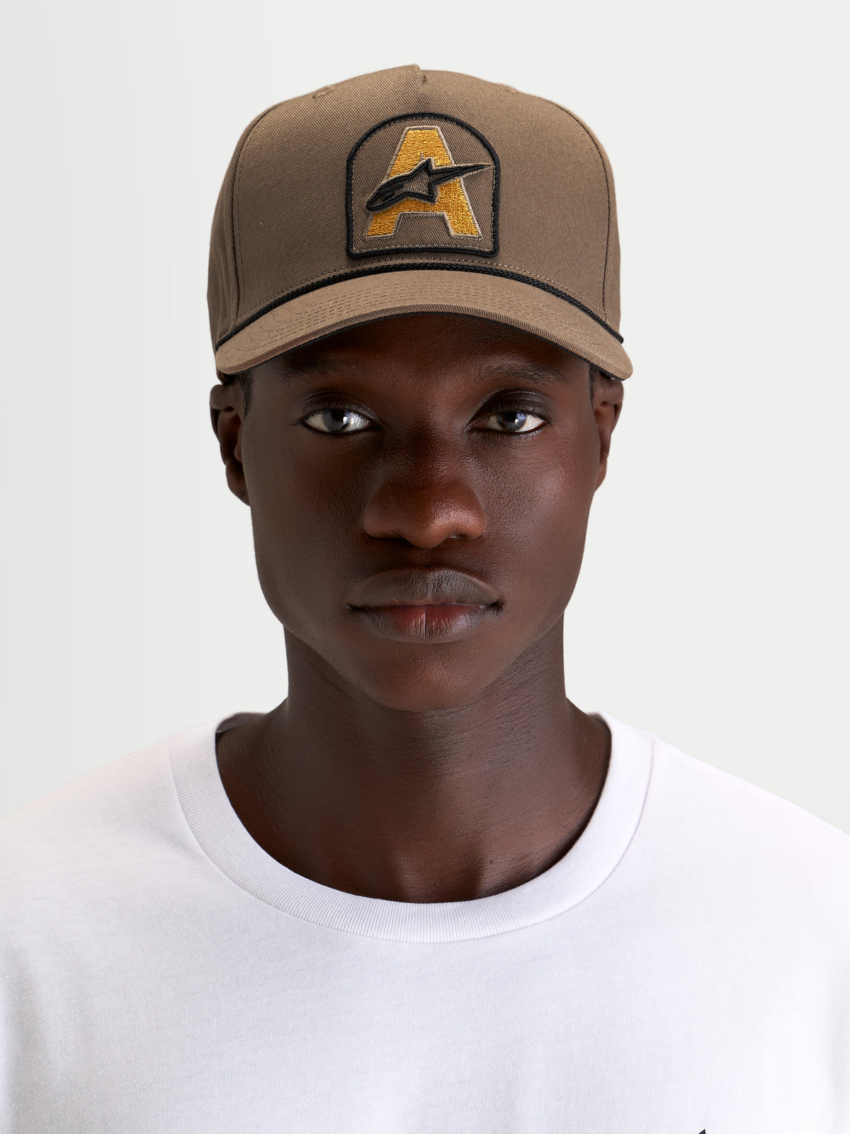 Alpinestars Czapka Acme Snapback, brązowa czapka z daszkiem w stylu sportswear, na modelu, z usztywnioną koroną, zakrzywionym daszkiem oraz przednią naszywką z haftowanym logo Astars i ozdobnym sznurkiem