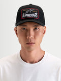 Alpinestars Hemmings Trucker czapka z daszkiem sportswear, czarna, z zakrzywionym daszkiem, oddychającym tyłem z siatki i tkaną naszywką z logo z przodu, prezentowana przez modela
