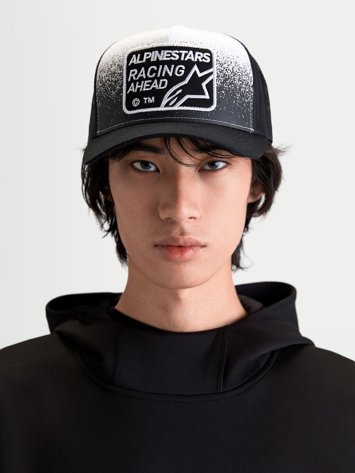Alpinestars czapka z daszkiem Gravel, lifestylowa czapka sportowa, czarna i biała, widok z przodu na modelu, zakrzywiony daszek, oddychający tył z siatki oraz tkana naszywka Racing Ahead z detalami o fakturze żwiru.