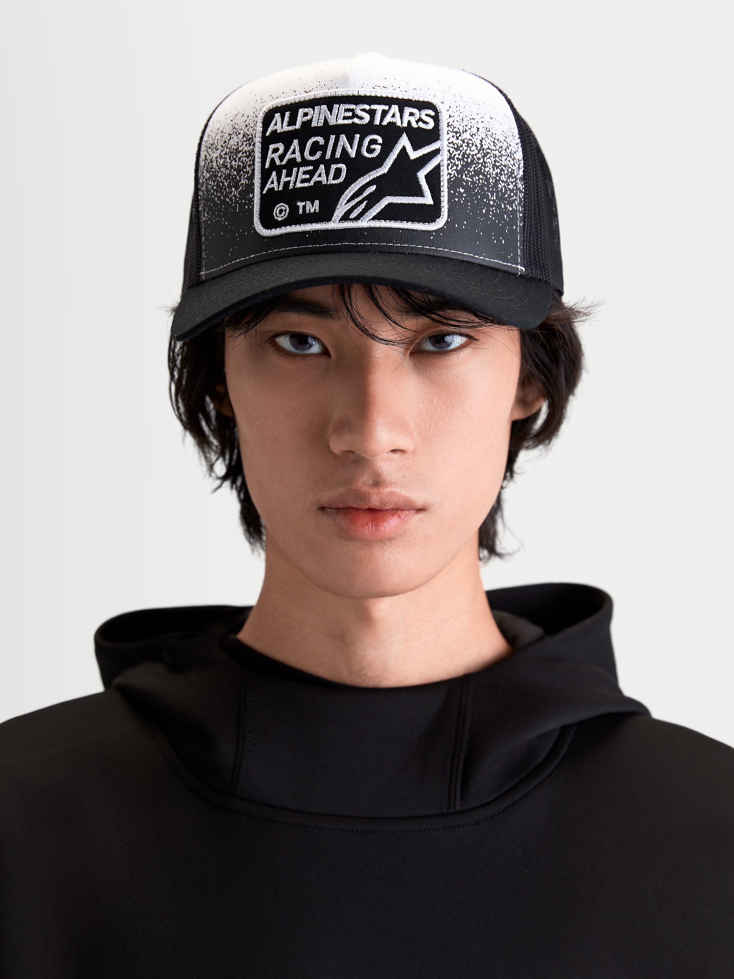 Alpinestars czapka z daszkiem Gravel, lifestylowa czapka sportowa, czarna i biała, widok z przodu na modelu, zakrzywiony daszek, oddychający tył z siatki oraz tkana naszywka Racing Ahead z detalami o fakturze żwiru.