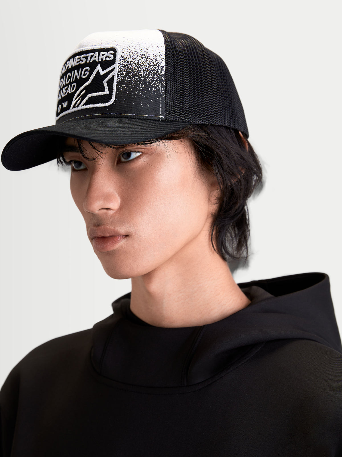Alpinestars Czapka Z Daszkiem Gravel, czapka z daszkiem typu trucker na modelu, czarna i biała, usztywniona korona, zakrzywiony daszek, przewiewny siatkowy tył, tkana naszywka z grafiką o teksturze żwiru i brandingiem Alpinestars Racing Ahead, casualowa odzież sportowa.