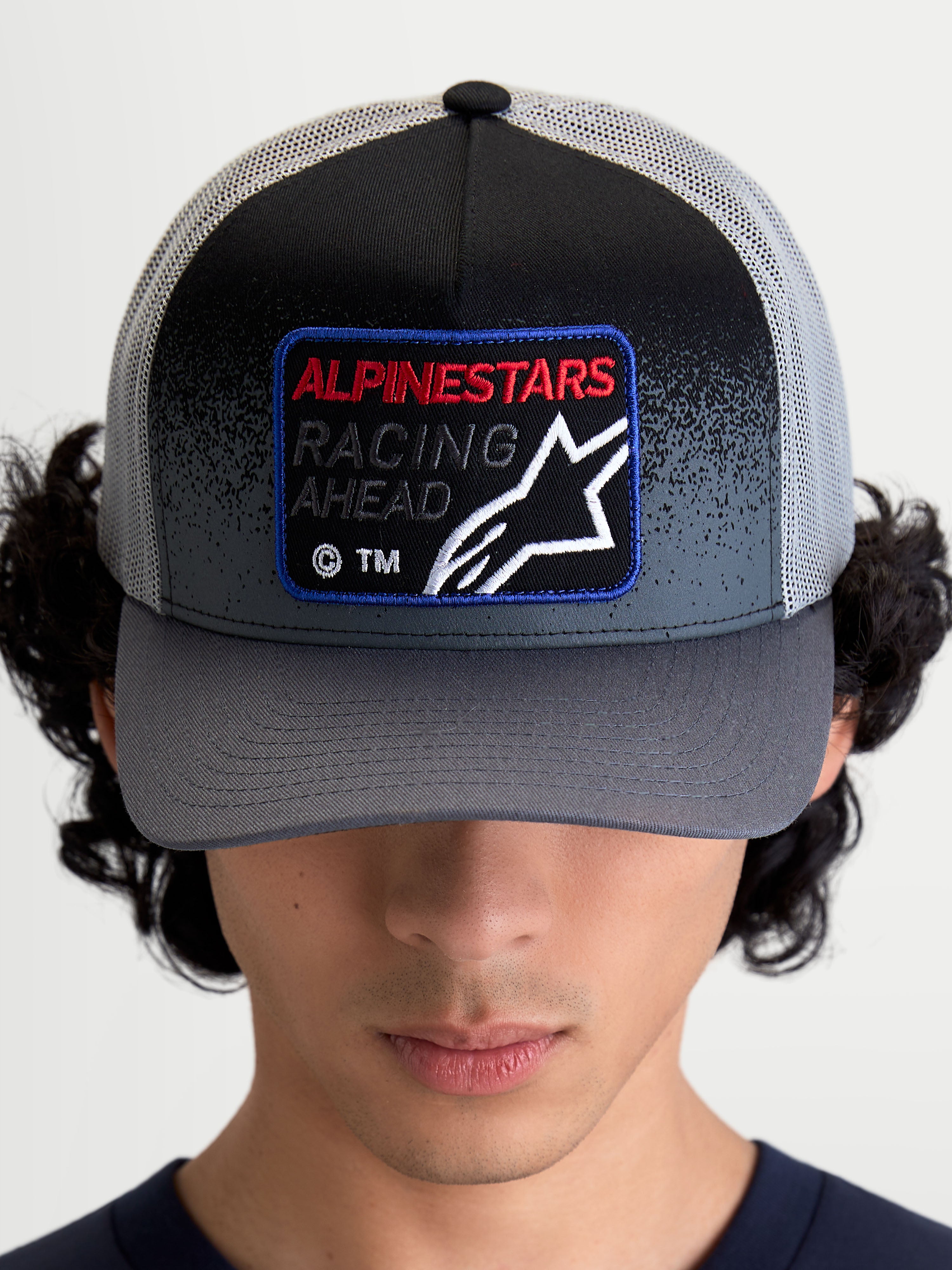 Alpinestars Czapka Z Daszkiem Gravel, sportowa czapka trucker, szara i czarna, tkana naszywka z przodu z napisem Racing Ahead i logo Astars, zakrzywiony daszek, tył z siatki, widok na modelu z przodu.