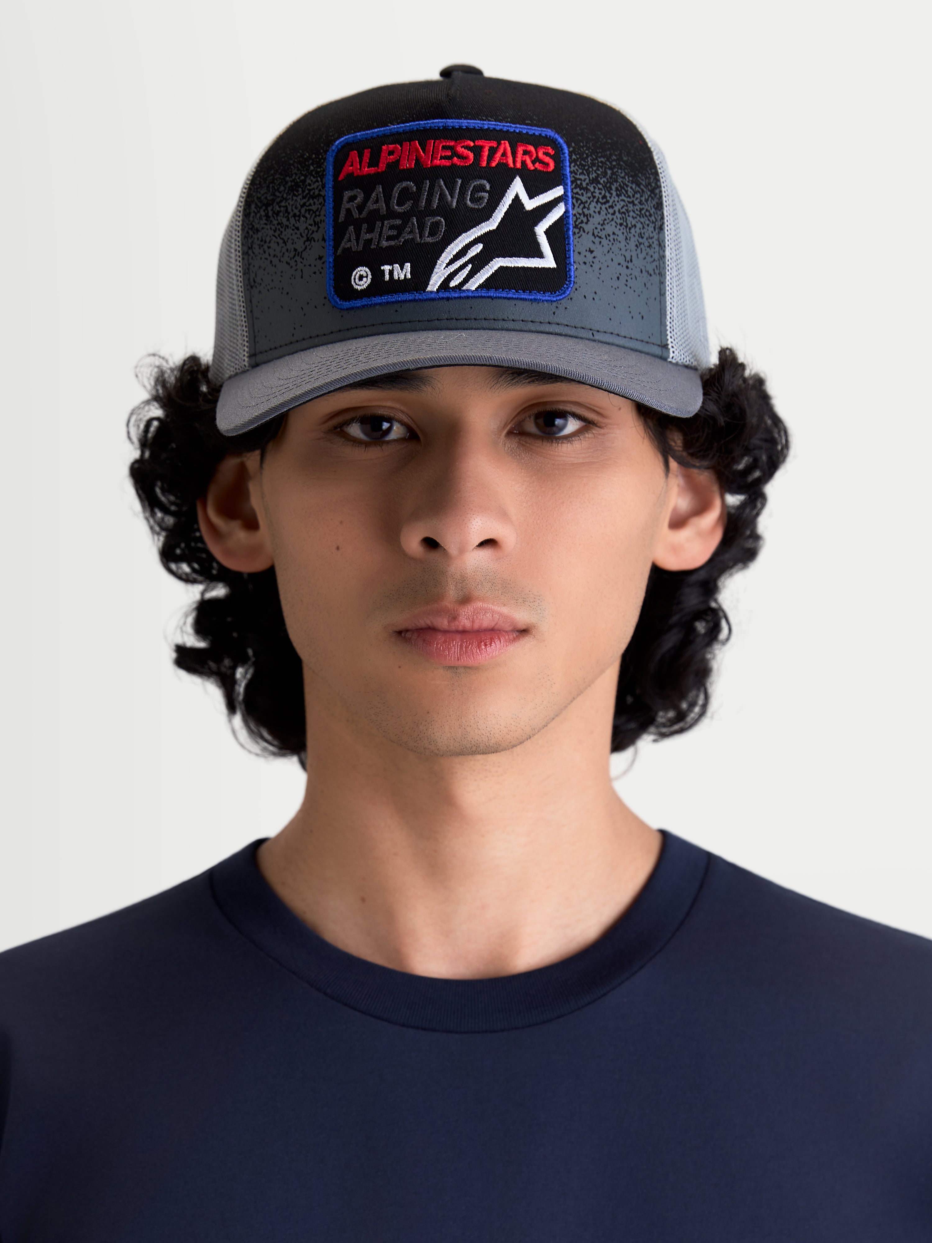 Alpinestars Gravel Trucker Hat, czapka z daszkiem, ciemnoszara i czarna, unisex, usztywniona korona, zakrzywiony daszek, tył z siatki, tkana naszywka Alpinestars Racing Ahead z detalami o teksturze żwiru, produkt na modelu.