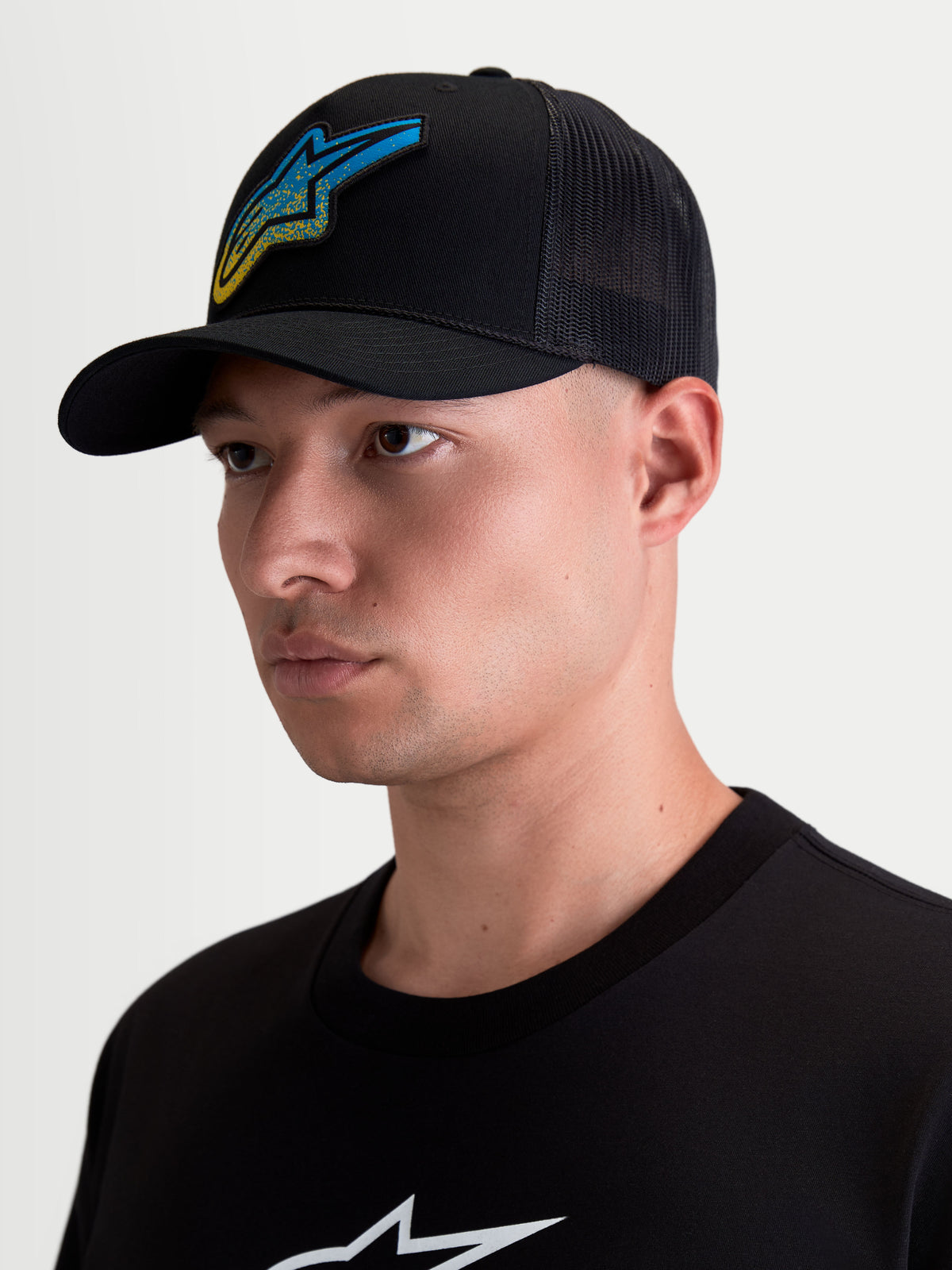 Alpinestars czapka z daszkiem Meteorite, męska czapka typu trucker, czarna, niebiesko-żółta tkana naszywka Astars o fakturze żwiru, zakrzywiony daszek i usztywniona korona, przewiewna siatka z tyłu, produkt prezentowany na modelu.