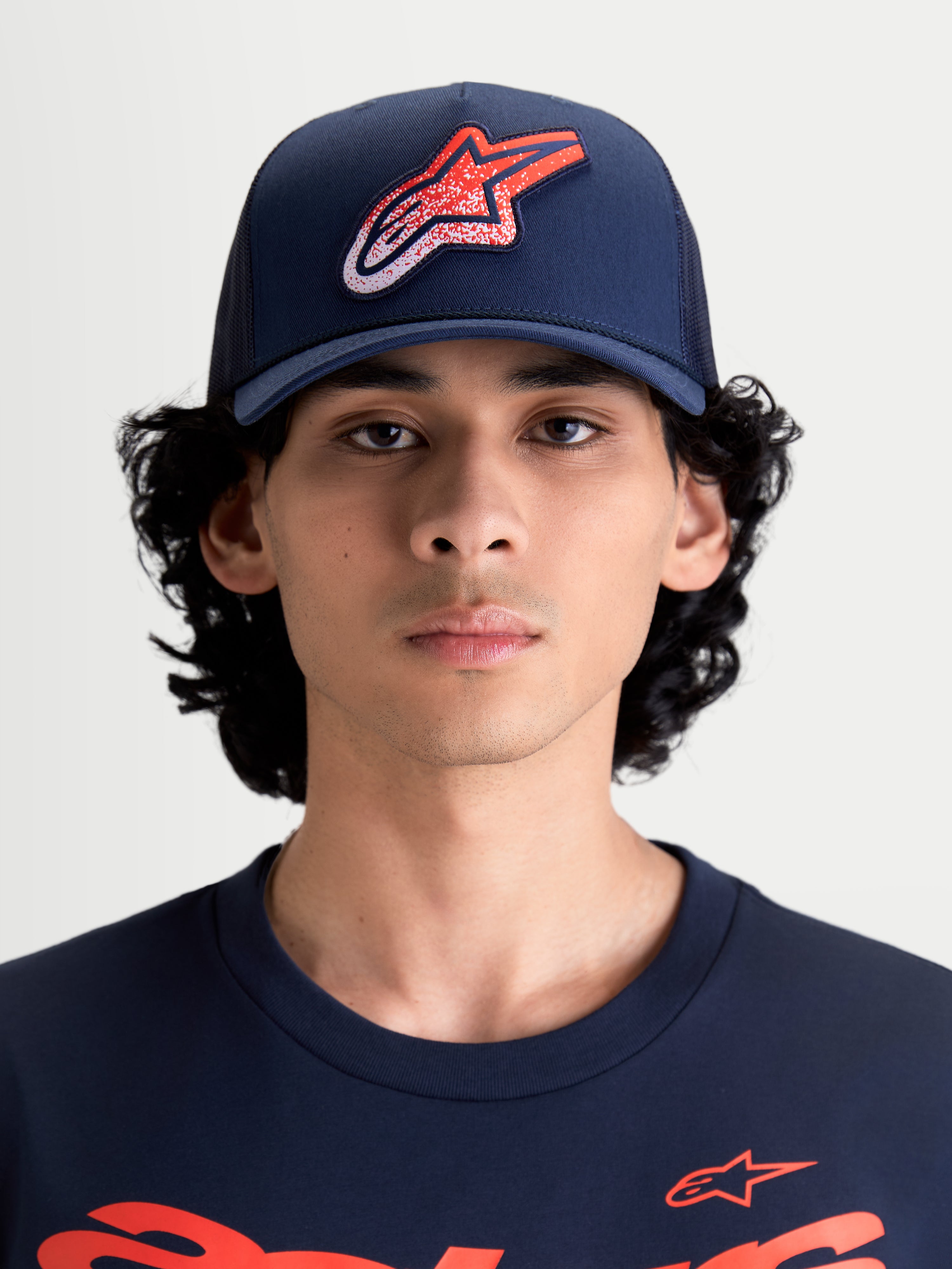Alpinestars Meteorite, czapka z daszkiem typu trucker, odzież sportowa dla kobiet i mężczyzn, granatowa z czerwono-białym logo Astars o teksturze żwiru, usztywniona korona, zakrzywiony daszek, tył z siatki, widok z przodu na modelu