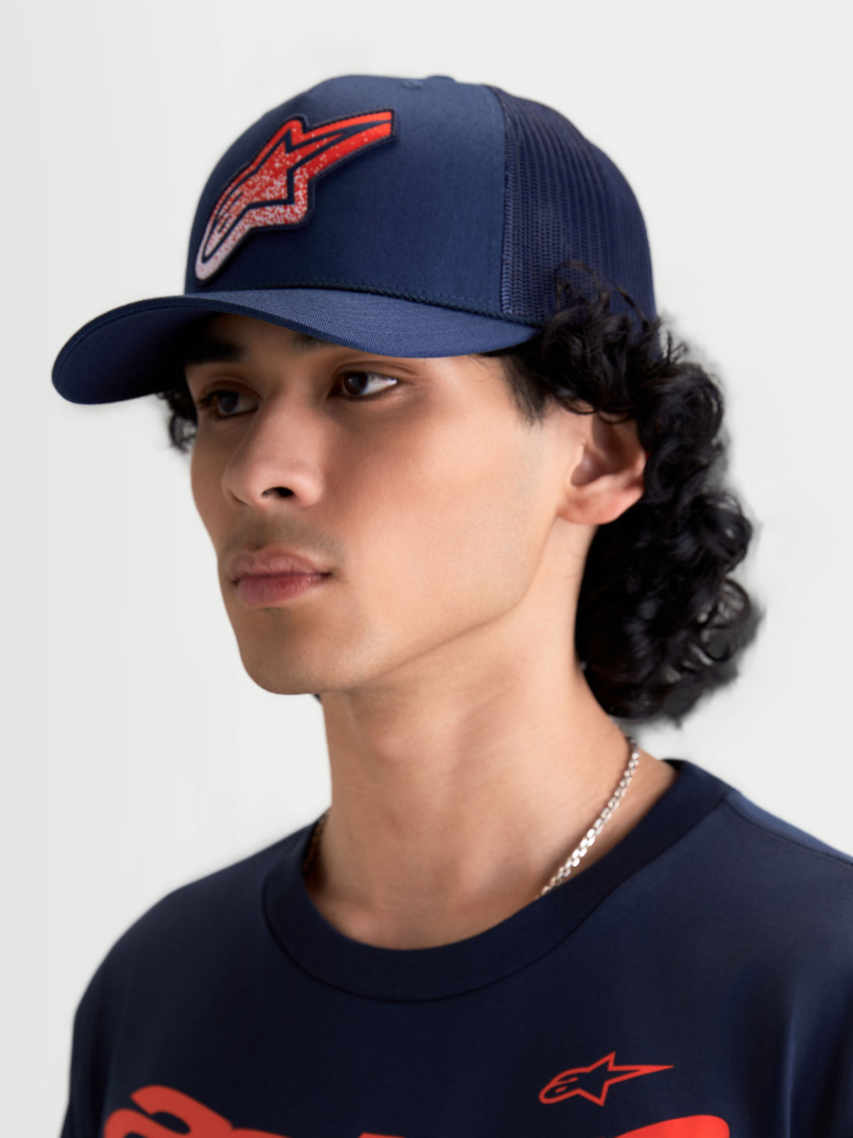 Alpinestars Czapka Z Daszkiem Meteorite, sportowa czapka z siatką typu trucker, granatowa z czerwonym logo Astars o teksturze żwiru, zakrzywiony daszek i usztywniona korona, widok na modelu.