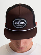 Alpinestars Czapka Z Daszkiem Hotlink, brązowa i czarna czapka typu trucker na modelu, usztywniona korona, płaski daszek z białym sznurkiem, naszywka z logo Astars Worldwide z obramowaniem w kształcie łańcucha rowerowego, odzież sportowa inspirowana motorsportem.