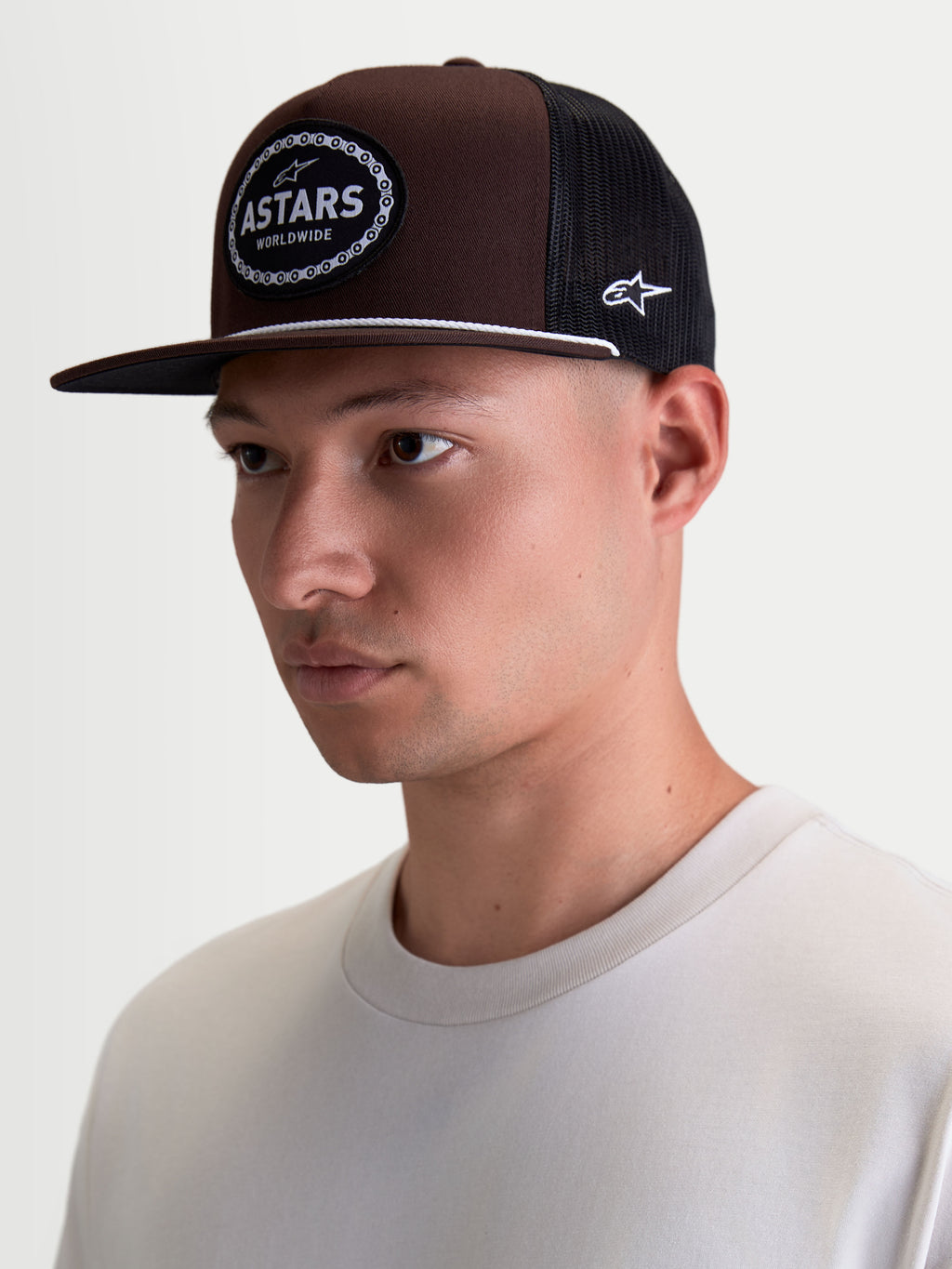 Alpinestars Czapka Z Daszkiem Hotlink, czapka typu trucker lifestyle na modelu, brązowa i czarna, płaski daszek, usztywniona korona z kepru, tkana naszywka z logo Astars i obramowaniem w kształcie łańcucha rowerowego.