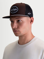 Alpinestars Czapka Z Daszkiem Hotlink, czapka typu trucker lifestyle na modelu, brązowa i czarna, płaski daszek, usztywniona korona z kepru, tkana naszywka z logo Astars i obramowaniem w kształcie łańcucha rowerowego.