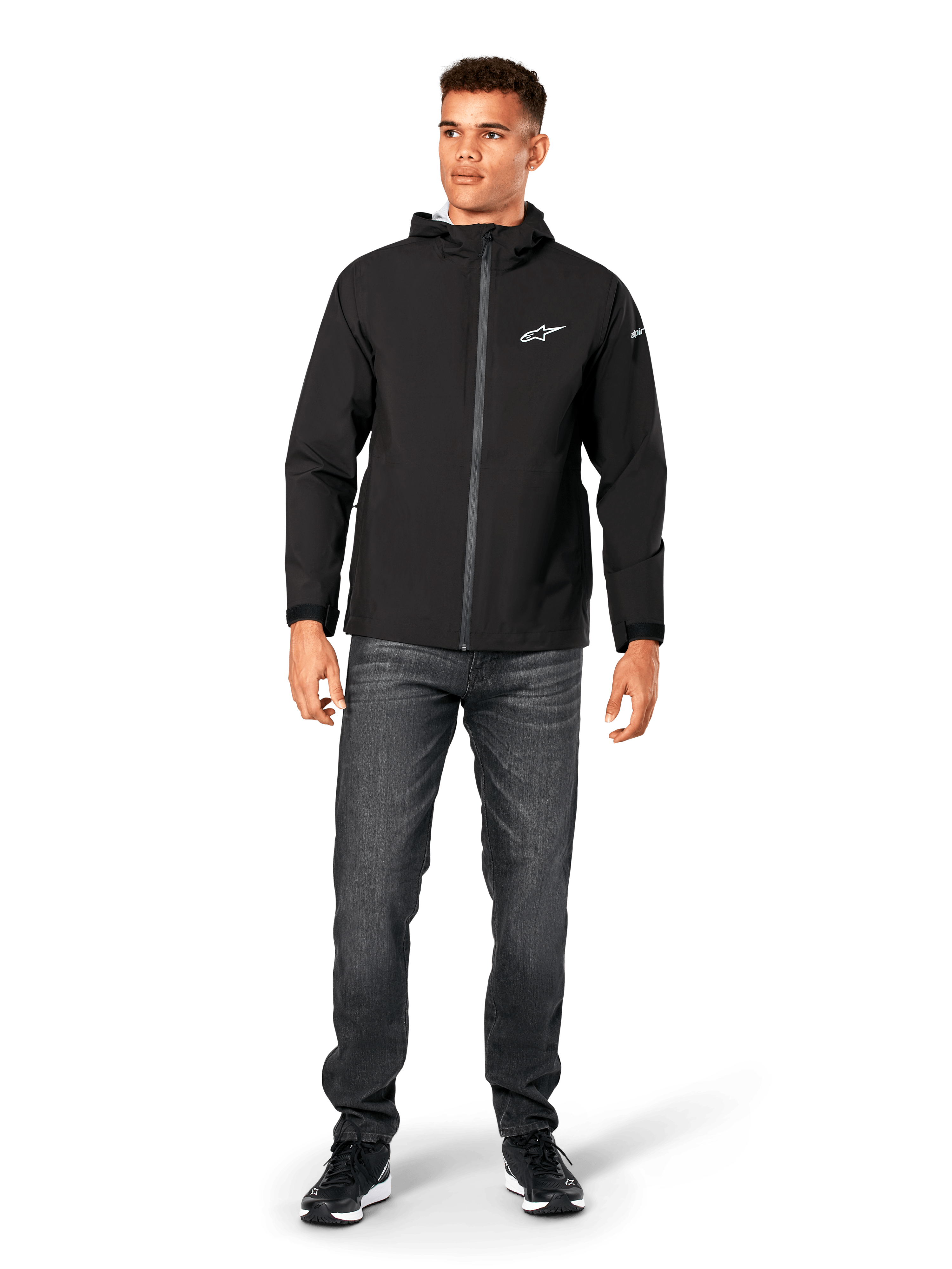 Kurtka przeciwdeszczowa Alpinestars Kascade, męska kurtka przeciwdeszczowa, czarna, noszona z ciemnoszarymi jeansami i czarnymi sneakersami, model z kapturem, powlekany zamek z przodu, białe logo Alpinestars na piersi, streetwear inspirowany motorsportem