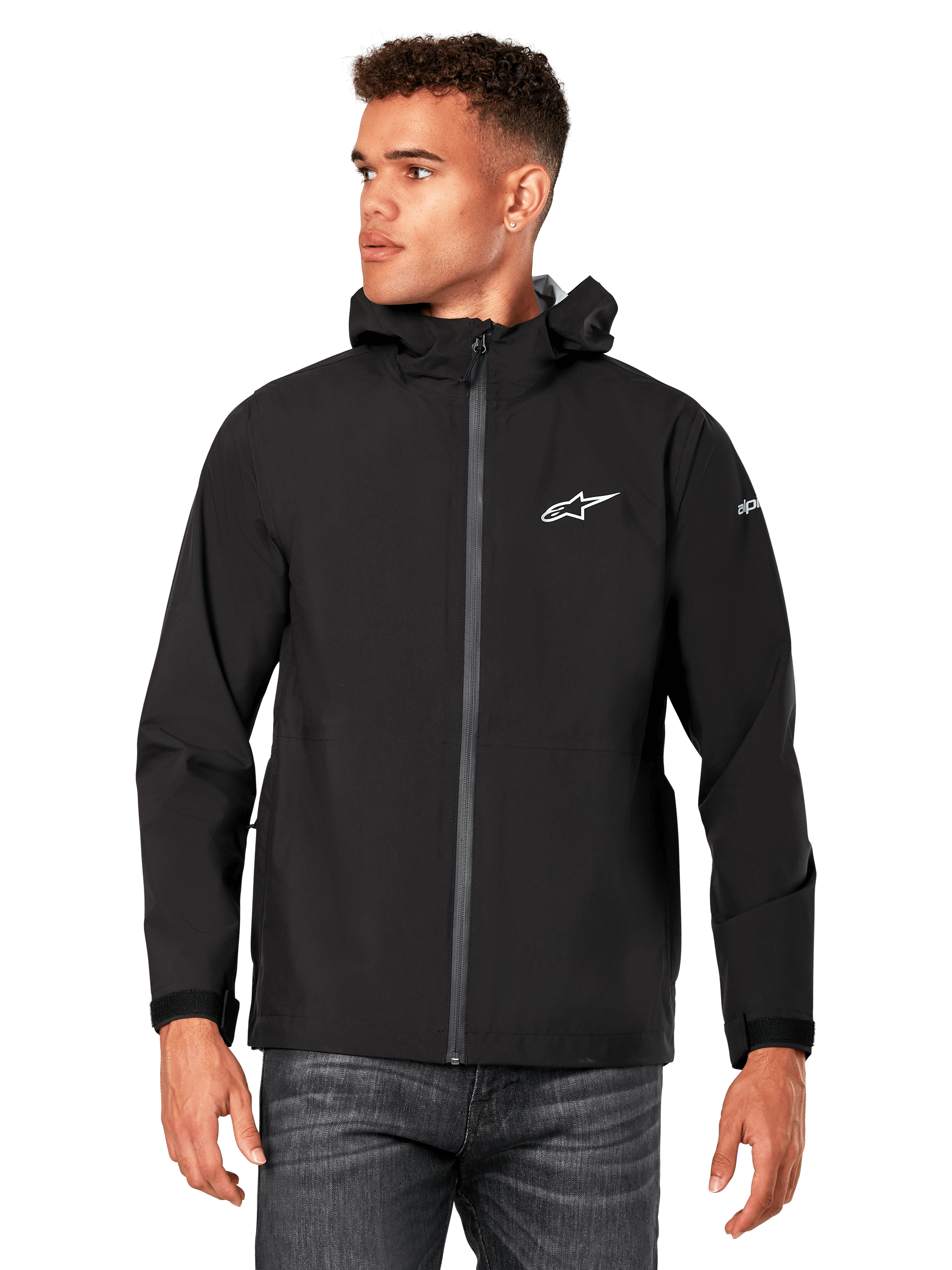 Mężczyzna w kurtce przeciwdeszczowej Alpinestars Kascade, czarna, białe logo na klatce piersiowej i branding na rękawie, fason z kapturem, powlekany zamek i mankiety na rzepy, w zestawieniu z ciemnymi dżinsami, casualowy styl streetwear inspirowany motorsportem