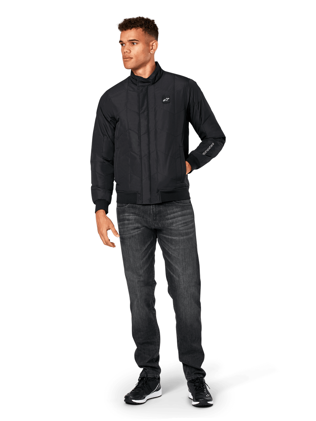 Kurtka Alpinestars Precedent, pikowana kurtka sportowa, czarna, noszona z ciemnoszarymi dżinsami i czarnymi sneakersami, ze skośnym wzorem pikowania, niskim kołnierzem, ściągaczami przy mankietach i na dole, białą naszywką z logo na piersi i haftem na rękawie