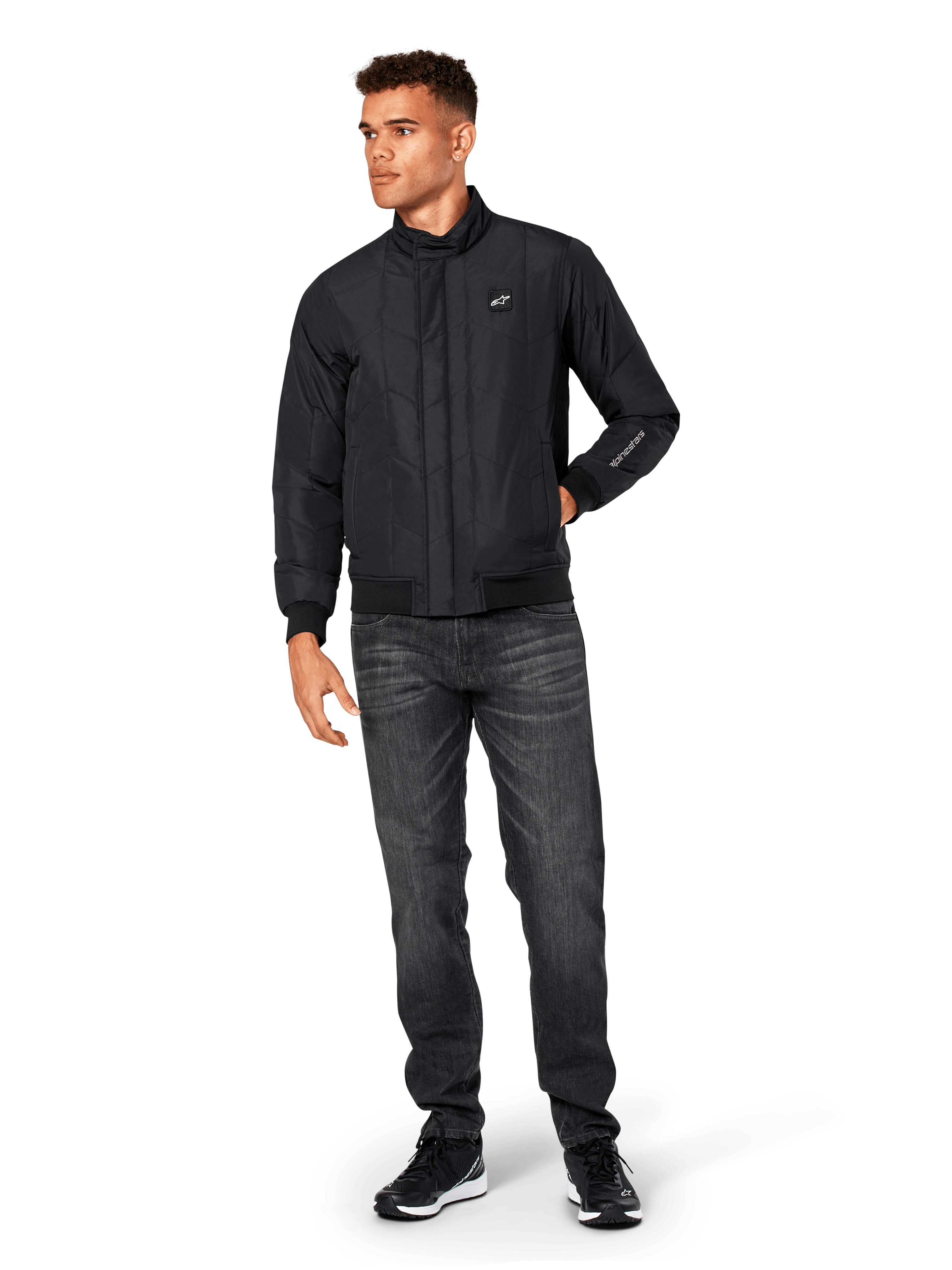 Kurtka Alpinestars Precedent, pikowana kurtka sportowa, czarna, noszona z ciemnoszarymi dżinsami i czarnymi sneakersami, ze skośnym wzorem pikowania, niskim kołnierzem, ściągaczami przy mankietach i na dole, białą naszywką z logo na piersi i haftem na rękawie