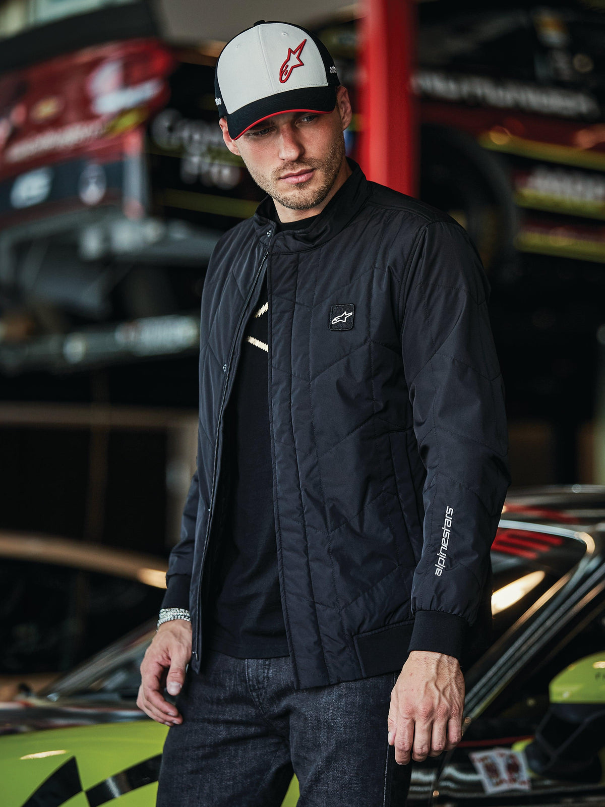 Alpinestars Precedent Jacket, czarna pikowana kurtka w stylu Formuły 1, z kultowym skośnym wzorem pikowania, naszywką z logo na piersi i białym haftem na rękawie, noszona z pasującą czapką Alpinestars w scenerii garażu sportów motorowych.
