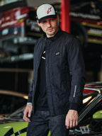 Alpinestars Precedent Jacket, czarna pikowana kurtka w stylu Formuły 1, z kultowym skośnym wzorem pikowania, naszywką z logo na piersi i białym haftem na rękawie, noszona z pasującą czapką Alpinestars w scenerii garażu sportów motorowych.