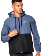 Męski model w kurtce Alpinestars Speeder Windbreaker Jacket, niebiesko-czarny design typu color-block, z kapturem i czarnymi sznurkami, białe logo na piersi, casualowa wiatrówka inspirowana sportami motorowymi