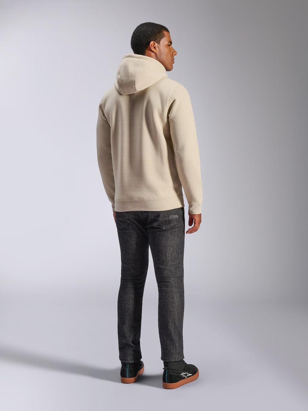 Alpinestars Ageless V3 Hoodie, codzienna polarowa bluza z kapturem w kolorze złamanej bieli widoczna od tyłu, prążkowane mankiety i dolny brzeg, noszona z ciemnymi dżinsami o kroju slim i czarnymi sneakersami Alpinestars