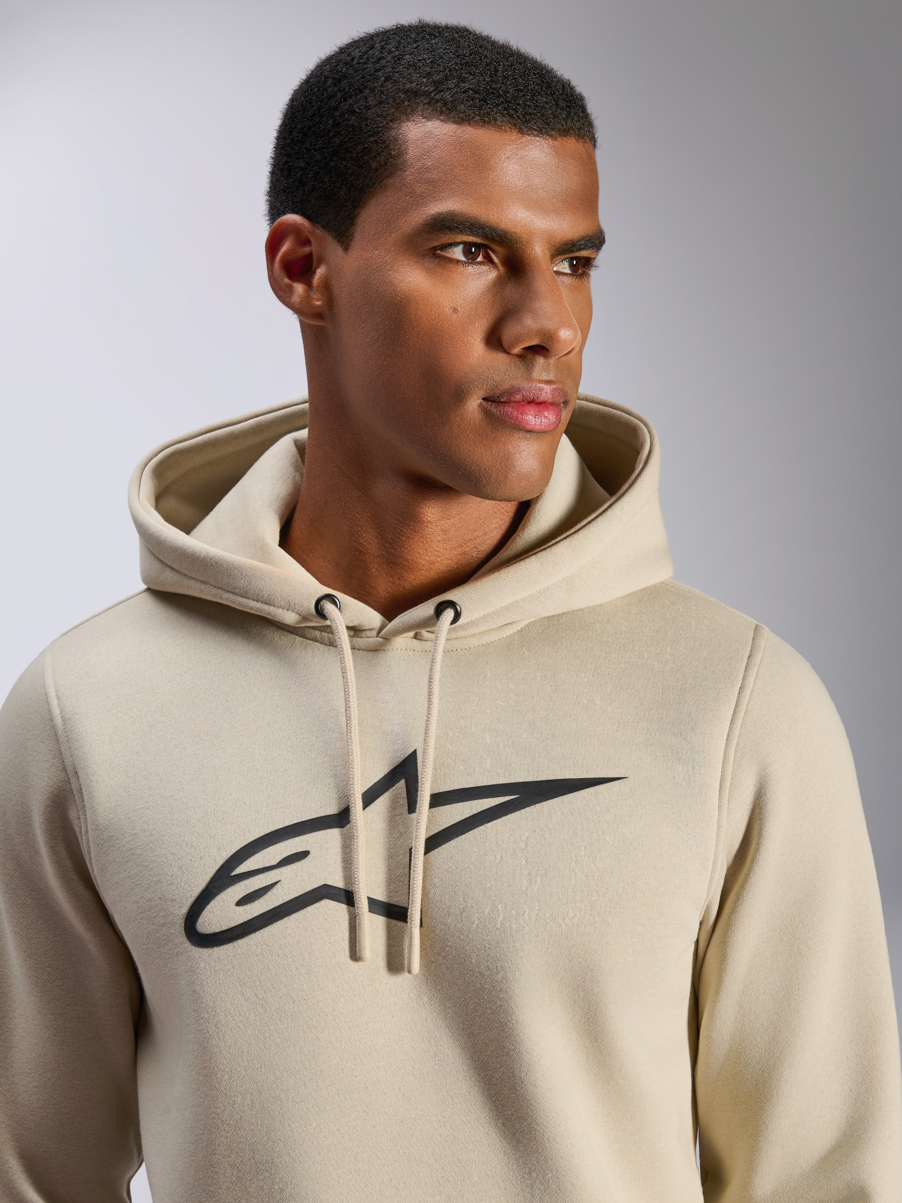 Alpinestars Ageless V3 Hoodie, sportowa odzież casual, złamana biel z czarnym haftowanym logo gwiazdy Alpinestars, kaptur ze sznurkiem, zbliżenie na modelu, miękki polar, nowoczesny design inspirowany motorsportem