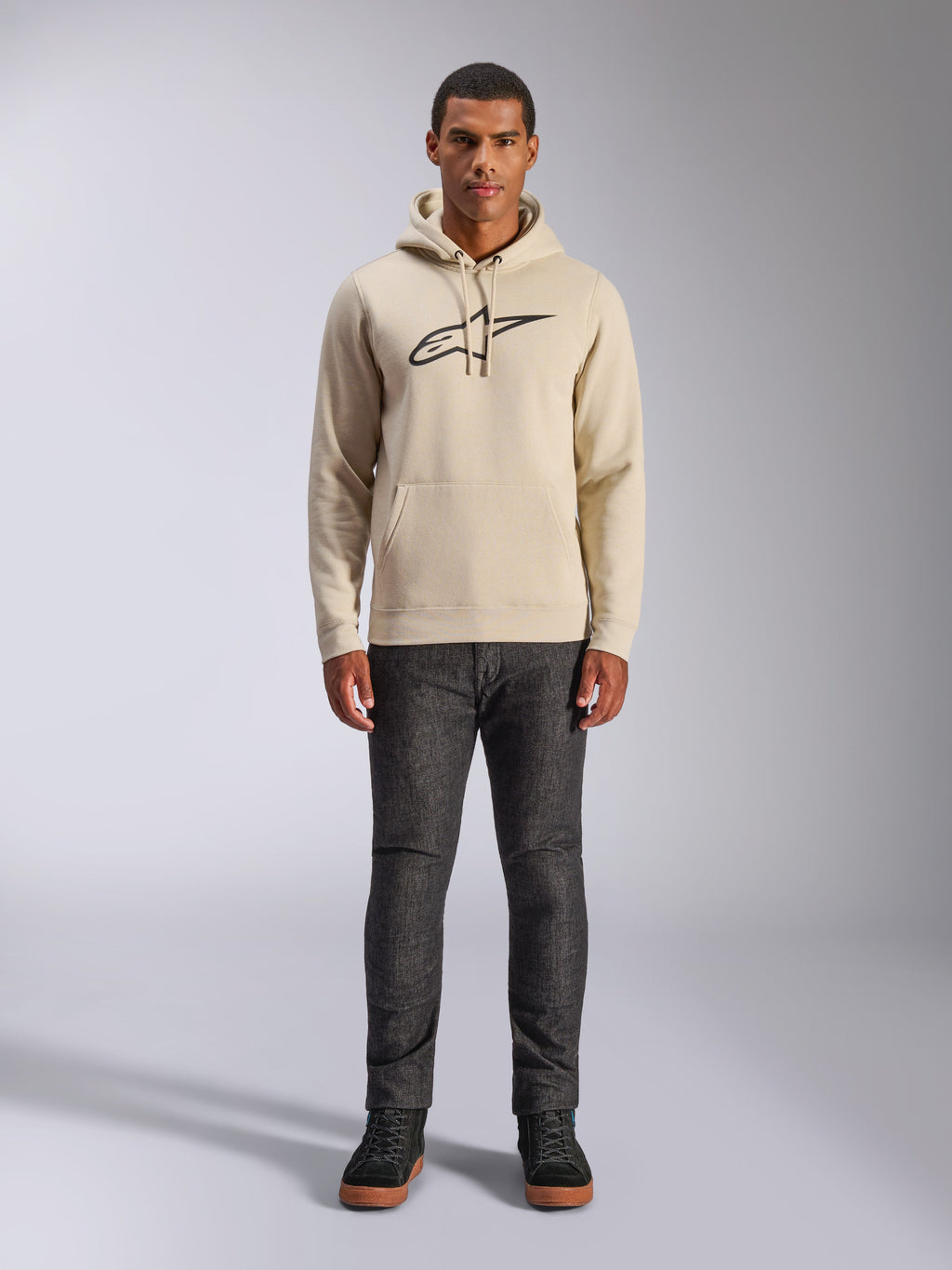 Alpinestars Ageless V3 Hoodie, codzienna odzież sportowa, złamana biel z czarnym haftowanym logo, noszona przez mężczyznę w ciemnoszarych spodniach i czarnych sneakersach, z kapturem ściąganym sznurkiem i kieszenią typu kangur, nowoczesny design inspirowany sportami motorowymi