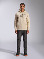 Alpinestars Ageless V3 Hoodie, codzienna odzież sportowa, złamana biel z czarnym haftowanym logo, noszona przez mężczyznę w ciemnoszarych spodniach i czarnych sneakersach, z kapturem ściąganym sznurkiem i kieszenią typu kangur, nowoczesny design inspirowany sportami motorowymi
