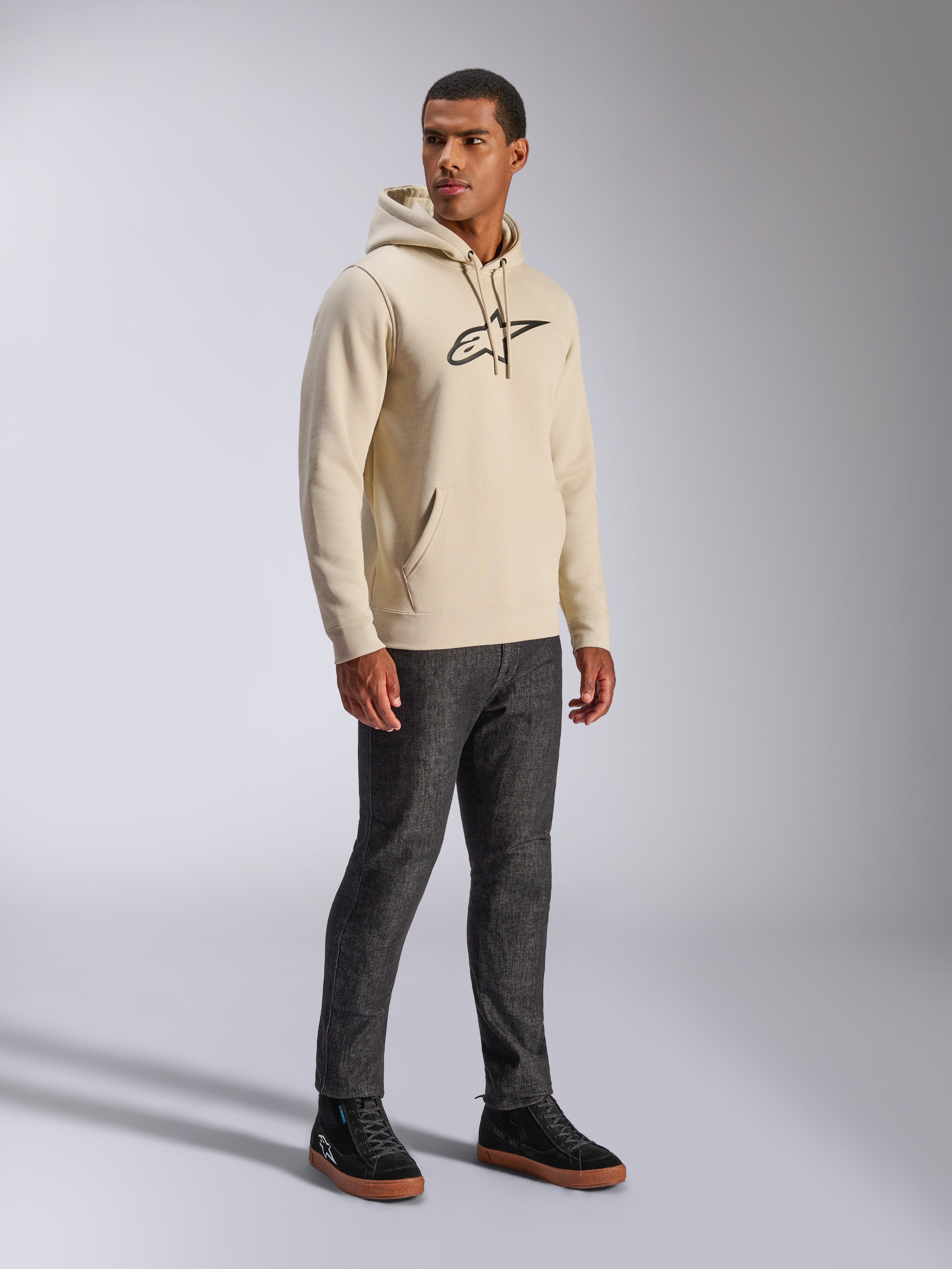 Alpinestars Ageless V3 Hoodie, codzienna odzież uliczna, kolor złamanej bieli z czarnym haftowanym logo, polarowa bluza z kapturem, długie rękawy, przednia kieszeń typu kangurka, zestawiona z ciemnymi dżinsami i czarnymi sneakersami