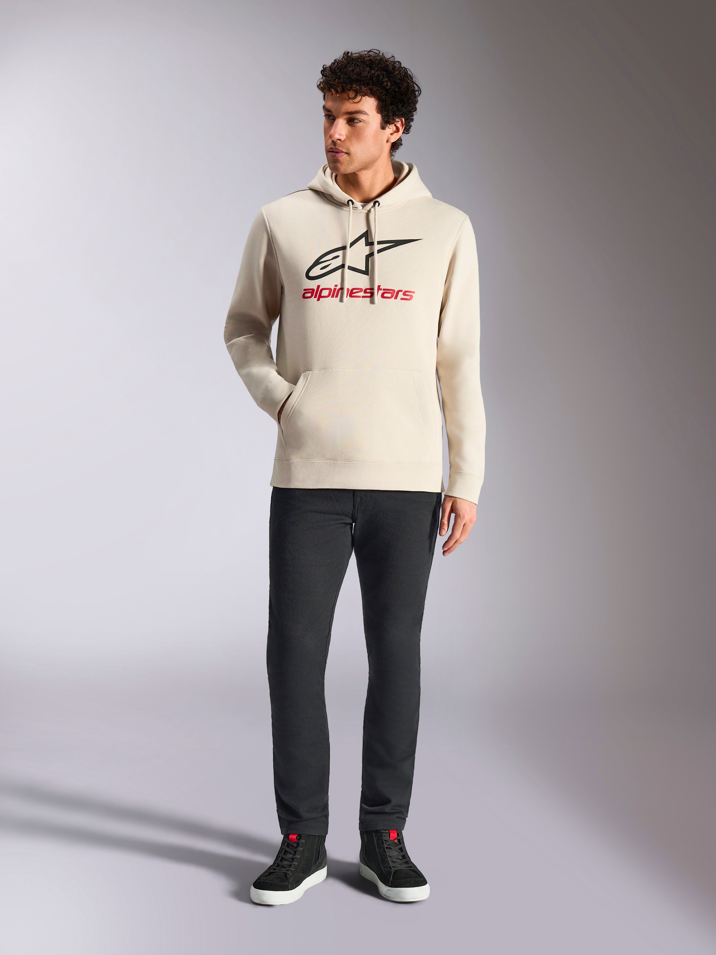 Alpinestars Always V3 Hoodie, casualowa odzież streetwear, kolor złamanej bieli z czarnym logo gwiazdy i czerwonym napisem, kaptur ze sznurkiem, długie rękawy, noszona przez modela z czarnymi spodniami i czarnymi sneakersami