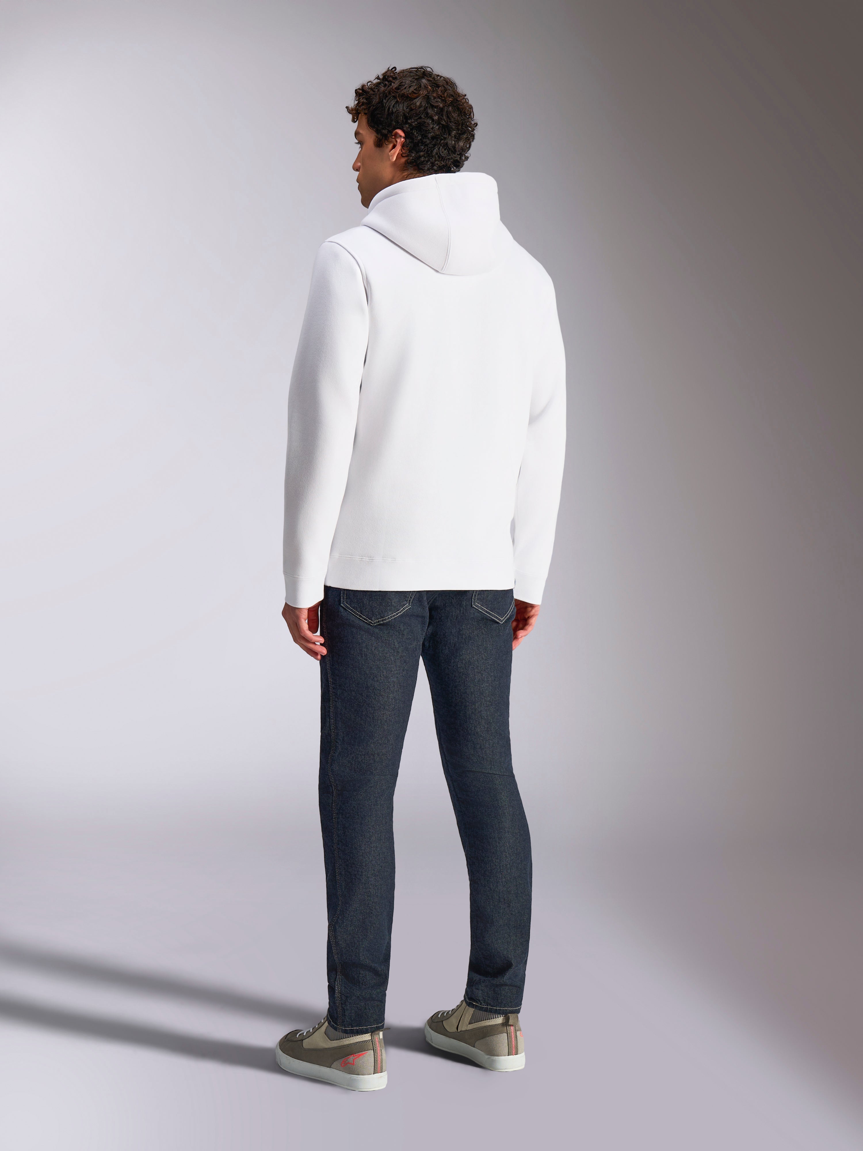 Alpinestars Blaze V3 Hoodie, widok z tyłu na modela w białej bluzie z kapturem, casualowa odzież sportowa zestawiona z ciemnoniebieskimi jeansami i szarymi sneakersami, nowoczesny design inspirowany sportami motorowymi