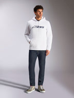 Alpinestars Blaze V3 Hoodie, biała codzienna bluza z kapturem typu streetwear z granatowym logo Alpinestars na klatce piersiowej, noszona przez modela z ciemnoniebieskimi jeansami i beżowymi sneakersami, wyposażona w kaptur ze sznurkiem i przednią kieszeń typu kangurka