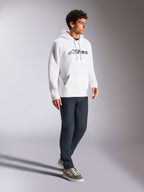 Bluza z kapturem Alpinestars Blaze V3, biało-granatowa, casualowy streetwear noszony przez modela z ciemnoniebieskimi dżinsami i butami sportowymi, posiada kaptur ze sznurkiem i wyrazisty nadruk logo z przodu, nowoczesny design inspirowany motorsportem
