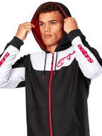 Alpinestars Sessions V3 Hoodie, czarno-biało-czerwona bluza rozpinana na zamek, noszona przez mężczyznę z założonym kapturem, czerwone logo Alpinestars na klatce piersiowej i rękawach, codzienna odzież motorsportowa typu streetwear
