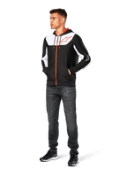 Alpinestars Sessions V3 Hoodie, codzienna bluza z kapturem na zamek, czarno-biało-czerwona, noszona przez modela z ciemnymi dżinsami i czarnymi sneakersami, białe panele color-block na klatce piersiowej i rękawach, czerwony zamek i detale sznurków, duże logo na piersi, polar poliestrowy, streetwear inspirowany motorsportem