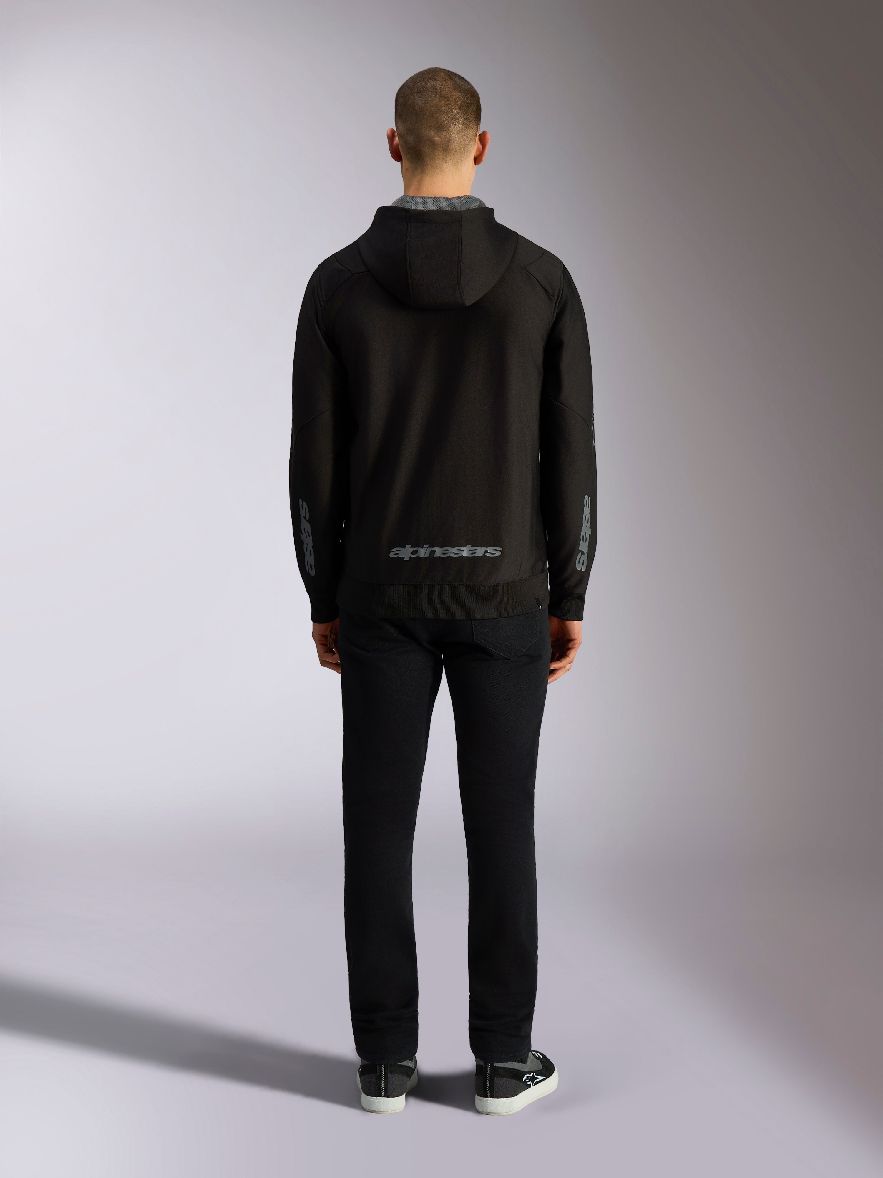 Alpinestars Sessions V3 Hoodie, czarno-szara bluza z kapturem, widok z tyłu, szary napis Alpinestars na dole pleców i logo astars na przedramionach, model w czarnych spodniach, codzienna odzież streetwearowa inspirowana sportami motorowymi