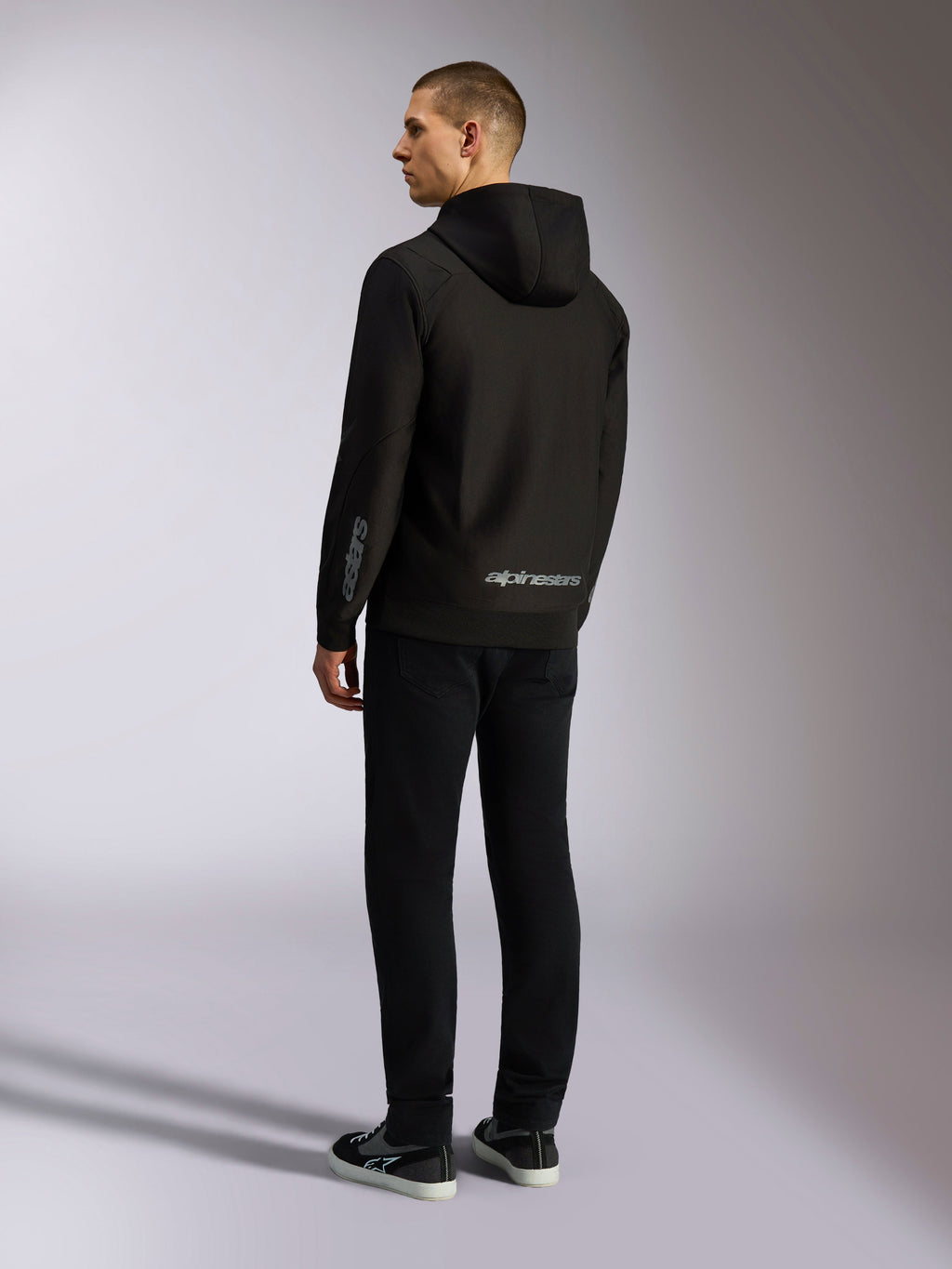 Alpinestars Sessions V3 Hoodie, czarna bluza streetwearowa, widok od tyłu na modelu, szary napis Alpinestars na dole pleców i logo Astars na rękawie, fason z kapturem zapinany na zamek, nowoczesny styl inspirowany motorsportem