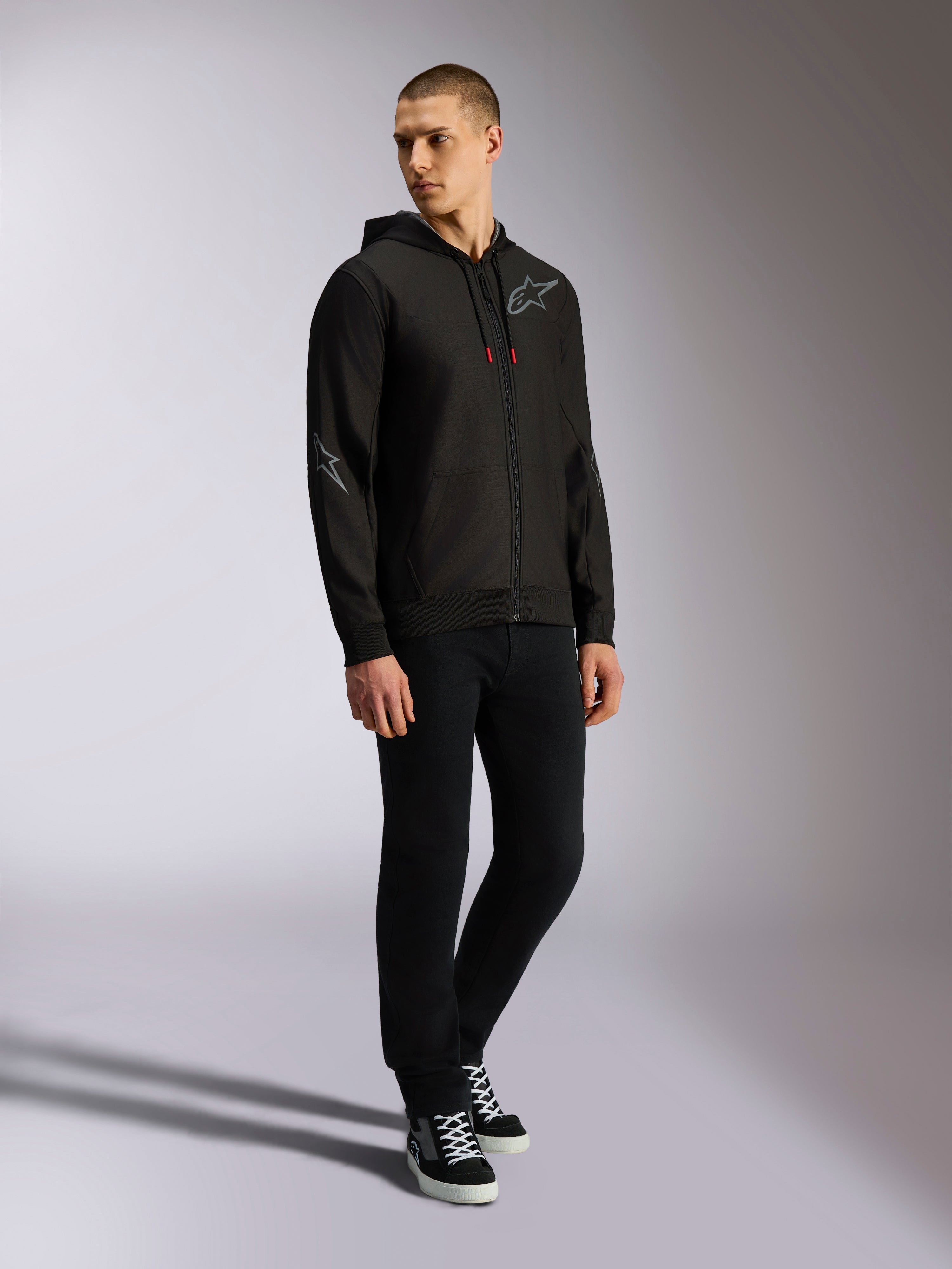 Alpinestars Sessions V3 Hoodie, czarno-szara polarowa bluza z zamkiem z przodu, szare logo na piersi i rękawach, kaptur ze sznurkami o czerwonych końcówkach, widok na całą sylwetkę modela w codziennym stroju streetwearowym inspirowanym motorsportem