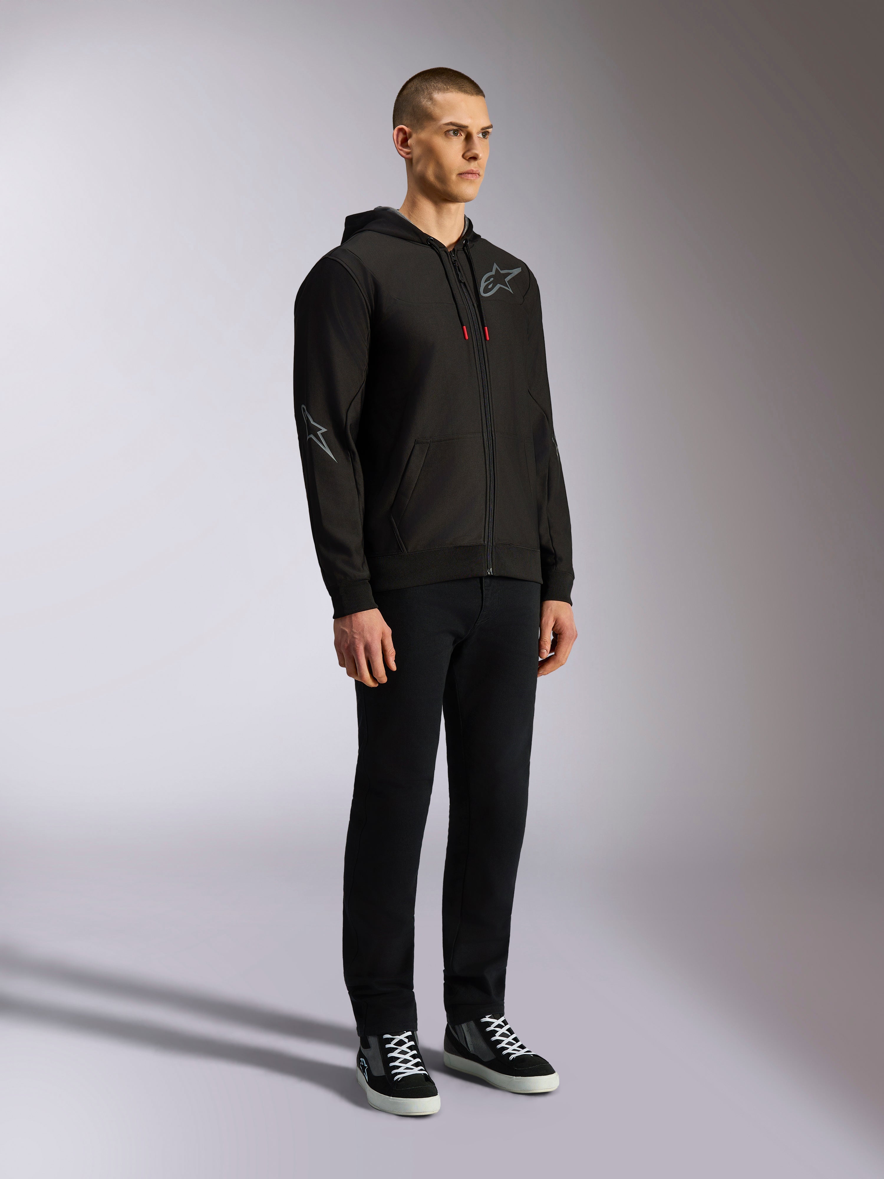 Bluza z kapturem Alpinestars Sessions V3, codzienna odzież streetwearowa w stylu motorsportowym, czarna bluza rozpinana z szarym logo gwiazdy na piersi i rękawie, sznurki z czerwonymi końcówkami, ściągacze na mankietach i dole, noszona przez modela z czarnymi spodniami i sneakersami.