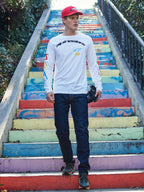 Alpinestars Brundo CSF Long Sleeve Tee, biała koszulka z długim rękawem, czarne logo Alpinestars i kolorowe grafiki, model na kolorowych schodach, ciemnoniebieskie dżinsy, czerwona czapka, codzienna odzież uliczna w stylu motorsport