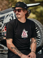 Czapka Alpinestars Gambit Trucker w kolorze czarnym z okrągłą naszywką z logo, noszona z czarną koszulką Alpinestars Soloist SS, zdjęcie lifestyle przedstawiające mężczyznę w okularach przeciwsłonecznych i z tatuażami siedzącego przed motocyklem motocrossowym.