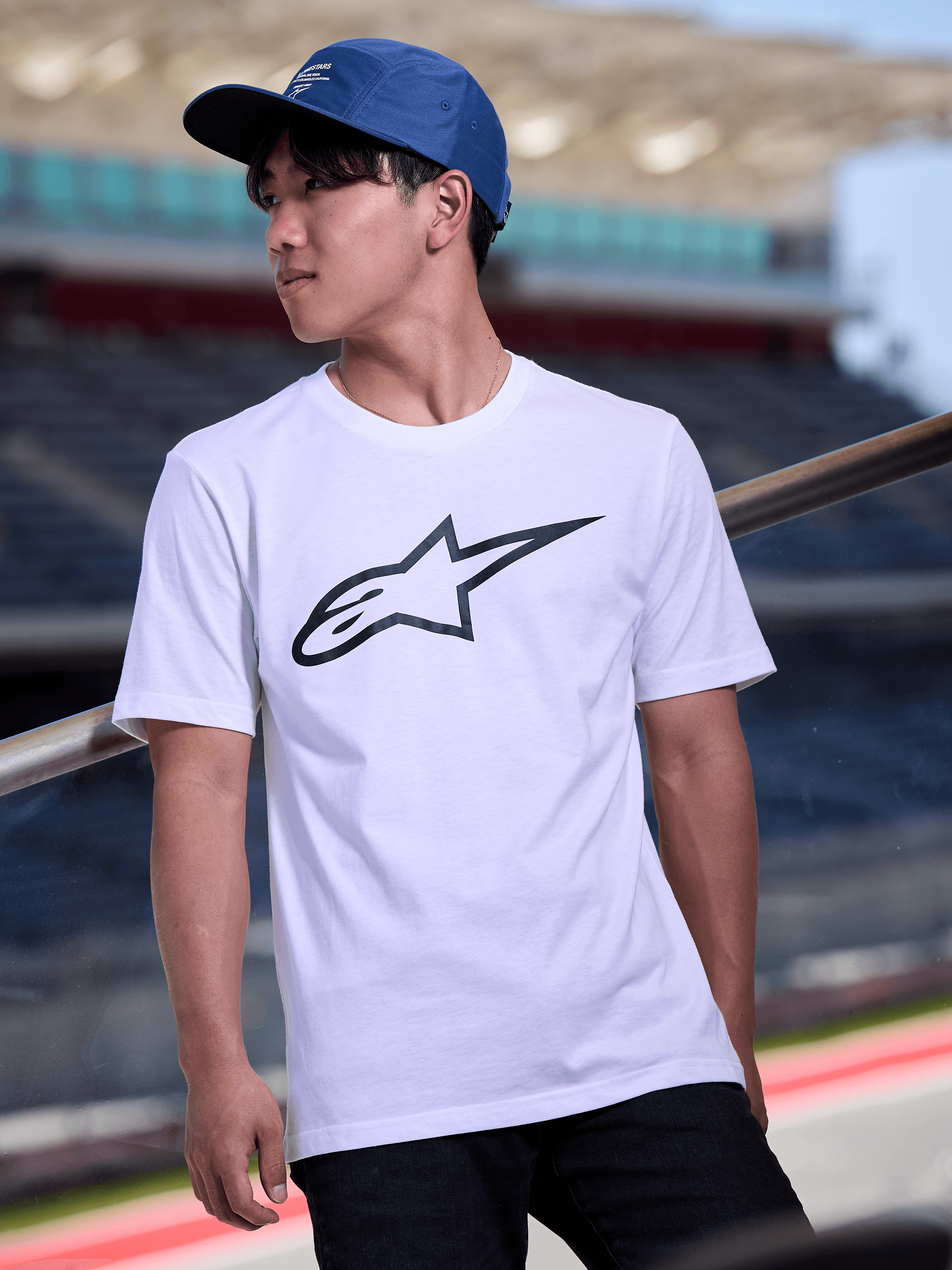 Mężczyzna w niebieskiej czapce Alpinestars Peasy Hat, 5-panelowa czapka typu jockey z płaskim daszkiem i białym nadrukiem graficznym, biały t-shirt z logo Alpinestars, streetwear inspirowany motorsportem