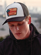 Alpinestars Hardware Trucker Hat w kolorze Military White, noszona przez mężczyznę w czarnej bluzie z kapturem, biały przedni panel, czarny zakrzywiony daszek i czarna siateczka z tyłu, beżowa haftowana naszywka z czerwonym brandingiem Alpinestars Motocross, casualowy styl lifestyle motorsport streetwear