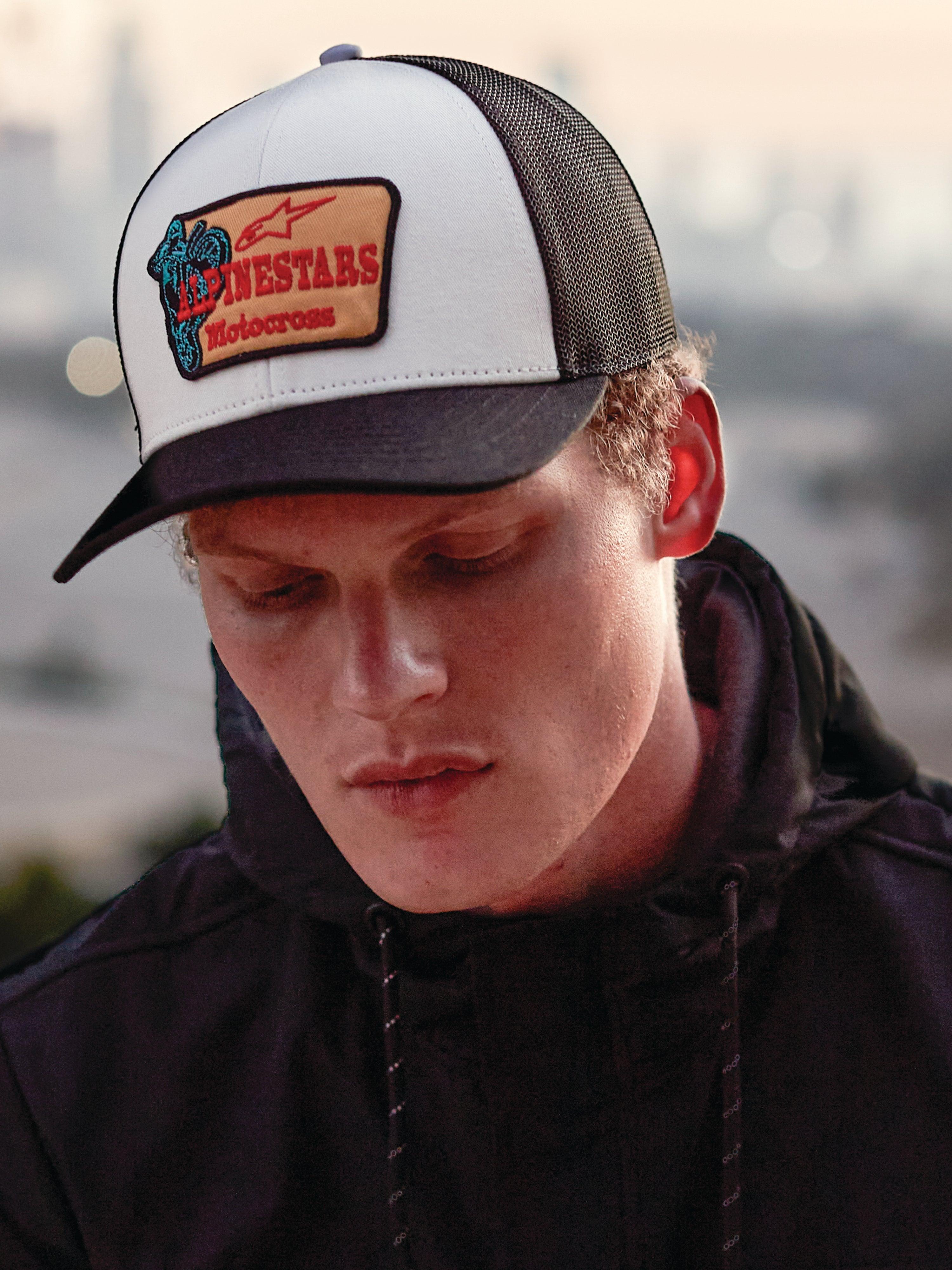 Alpinestars Hardware Trucker Hat w kolorze Military White, noszona przez mężczyznę w czarnej bluzie z kapturem, biały przedni panel, czarny zakrzywiony daszek i czarna siateczka z tyłu, beżowa haftowana naszywka z czerwonym brandingiem Alpinestars Motocross, casualowy styl lifestyle motorsport streetwear