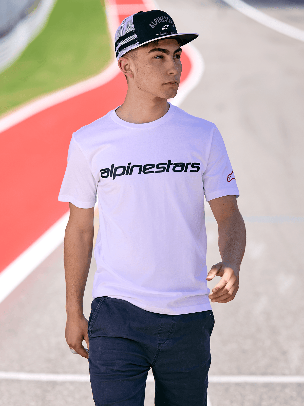Mężczyzna w czapce Alpinestars Backline Trucker Hat w kolorze czarno-białym, zestawionej z białym t-shirtem z logo Alpinestars i ciemnoniebieskimi spodniami, casualowy styl motorsportowy na tle toru wyścigowego