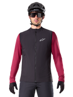Kamizelka Alpinestars A-Dura Thermal, czarna ocieplana kamizelka rowerowa, na modelu z bordową koszulką z długim rękawem, czarnym kaskiem MTB i okularami przeciwsłonecznymi, białe logo na piersi, lekka ochrona termiczna do jazdy na rowerze górskim