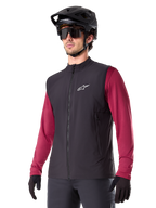 Alpinestars A-Dura Thermal Vest, czarna rowerowa kamizelka termiczna, noszona na bordowej koszulce z długim rękawem, z białym logo na piersi, w zestawie z czarnym kaskiem MTB i okularami przeciwsłonecznymi, izolująca warstwa techniczna do kolarstwa górskiego
