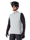 Kamizelka termiczna Alpinestars A-Dura, Titanium Gray, rowerowa kamizelka ocieplana, założona na czarną koszulkę z długim rękawem, z czarnym kaskiem MTB i rękawiczkami, różowe logo na piersi, pionowe boczne kieszenie na zamek