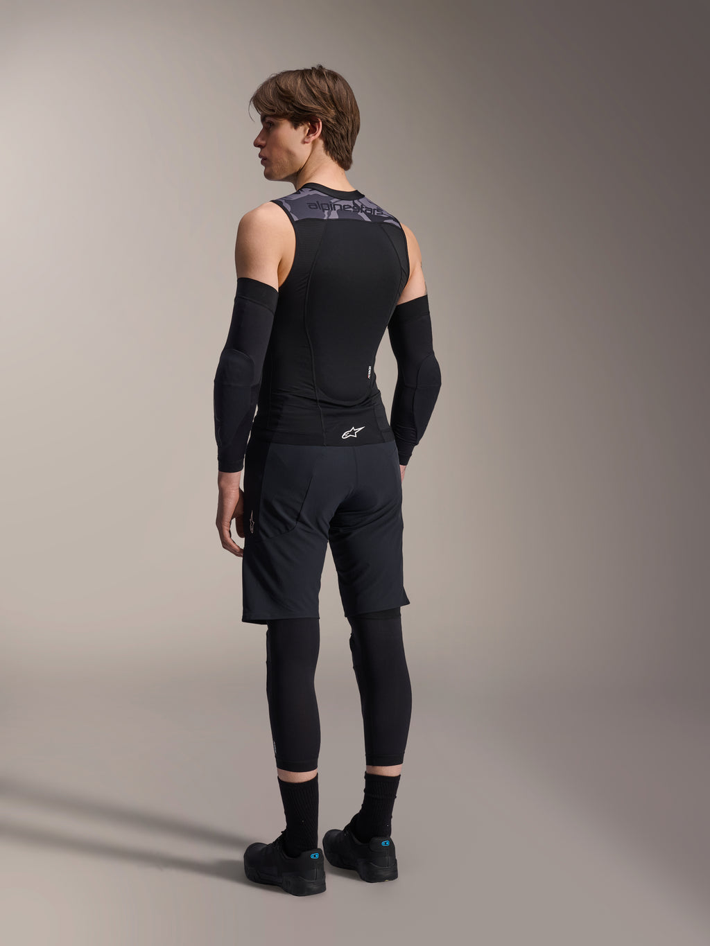 Męski model widoczny od tyłu w kamizelce ochronnej Alpinestars A-Motion Plasma Protection Vest, Black Camo, siateczkowy bezrękawnik z zintegrowanym ochraniaczem pleców, szary wzór kamuflażu na ramionach, czarne ochraniacze łokci i spodenki MTB, profesjonalna odzież rowerowa
