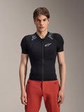 Męski model w kurtce ochronnej Alpinestars A-Motion Plasma z krótkim rękawem, czarna z szarym wzorem moro na ramionach, białe logo Alpinestars na klatce piersiowej, czerwone spodnie, dopasowana zbroja MTB z zamkiem z przodu