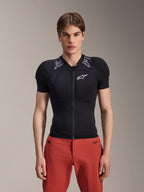 Męski model w kurtce ochronnej Alpinestars A-Motion Plasma z krótkim rękawem, czarna z szarym wzorem moro na ramionach, białe logo Alpinestars na klatce piersiowej, czerwone spodnie, dopasowana zbroja MTB z zamkiem z przodu