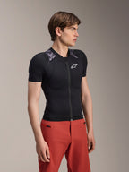 Męski model w kurtce ochronnej Alpinestars A-Motion Plasma Protection Jacket z krótkim rękawem, kolor Black Camo z szarymi panelami kamuflażowymi na ramionach, zintegrowane ochraniacze ramion i klatki piersiowej, białe logo gwiazdy na piersi, oddychający materiał siatkowy, zestawiona z czerwonymi spodniami